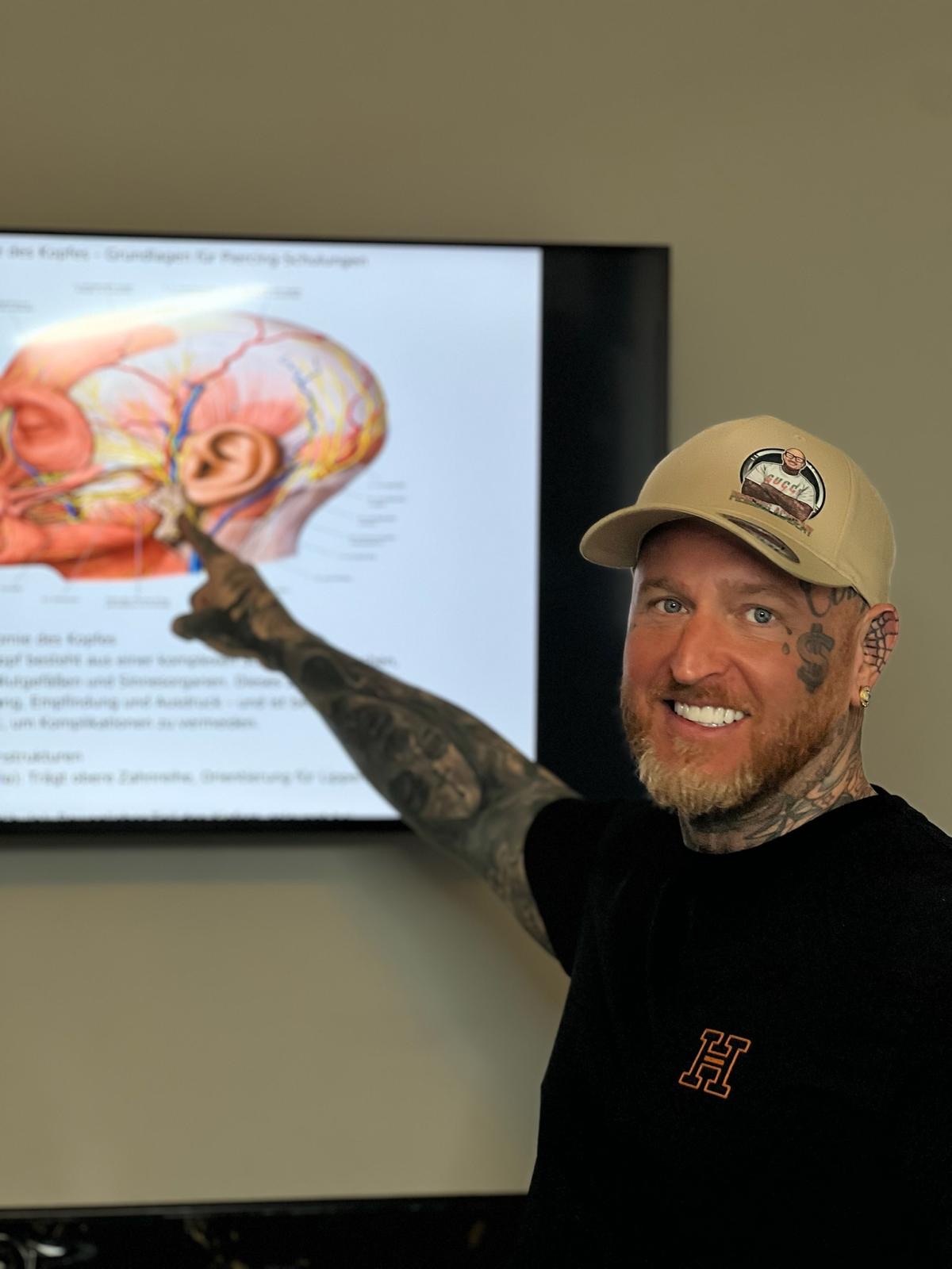 Tattoo Andy erklärt die Kopf-Anatomie vor einem Bildschirm in der Piercing-Schulung